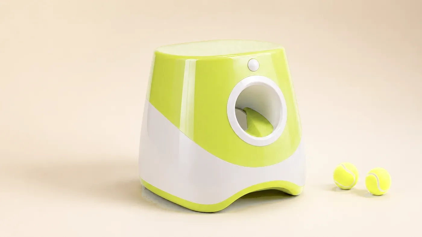 The yellow-green and white automatic ball launcher, seen three-quarters on a pale background, two balls beside it